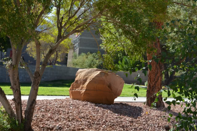 A large rock surrounded by trees and shrubs on a landscaped area with a grassy lawn and a building in the background. at UNLV