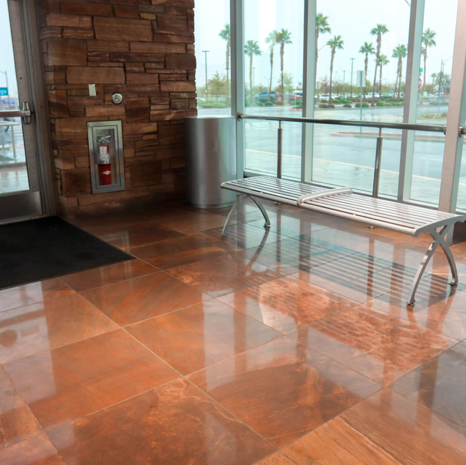 Lobby area with polished brown tile flooring, a glass and metal bench, large windows showing palm trees outside, and a door with a black mat.