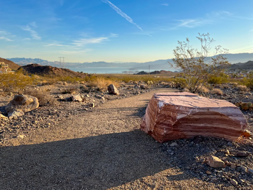 A desert landscape with a dirt path, a large striped rock on the right, scattered shrubs, and distant mountains under a blue sky.