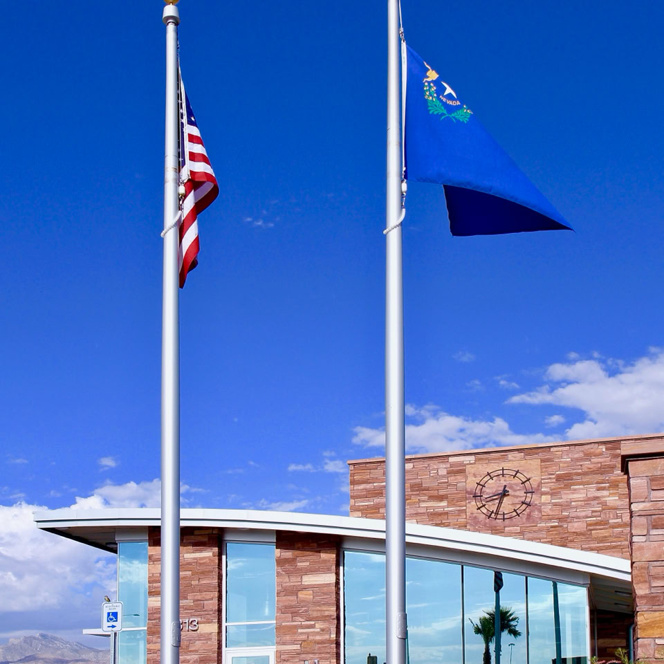 Two flagpoles with the American flag and a blue flag in front of a modern building with large glass windows, set against a clear blue sky.
