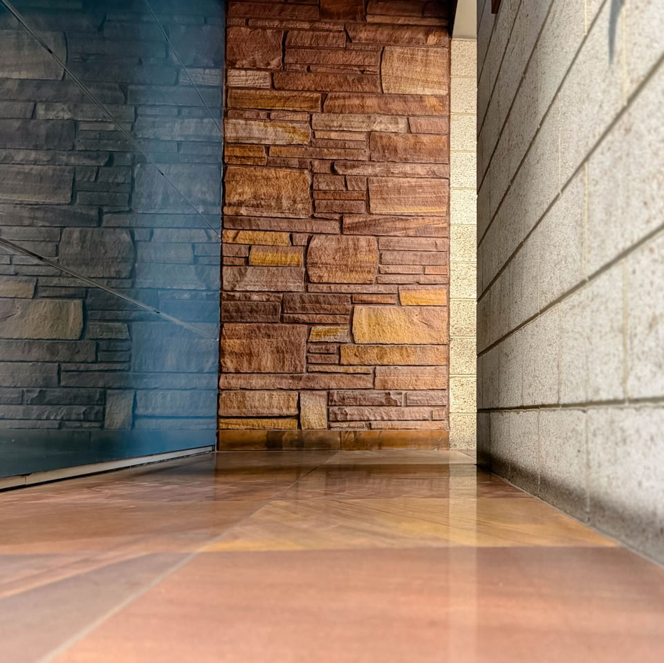 A hallway with a polished floor, a glass wall on the left, a brick wall at the end, and a concrete wall on the right.