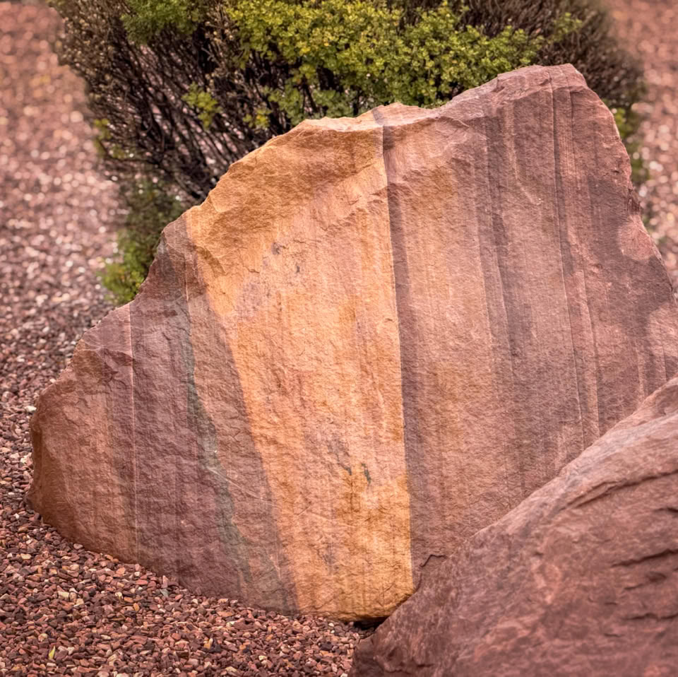 A large, textured rock with vertical stripes, surrounded by gravel and some greenery in the background.