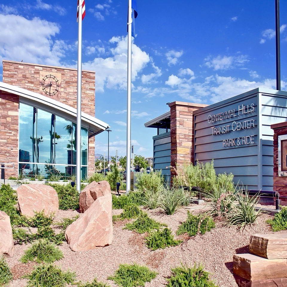 A modern building with large glass windows and stone accents, surrounded by desert landscaping with rocks and shrubs, under a clear blue sky.