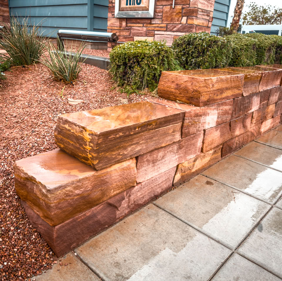 Stone retaining wall with large rectangular blocks, adjacent to a paved walkway and landscaped with shrubs and gravel.