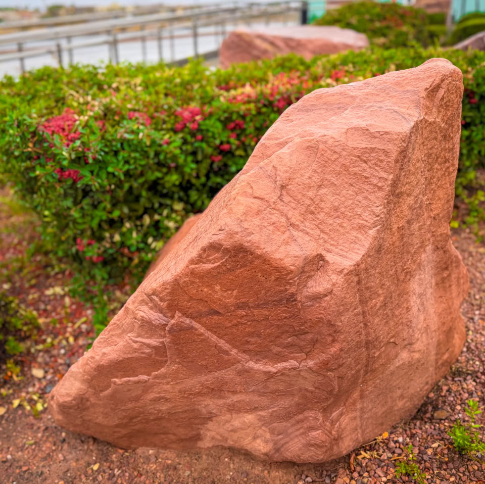 A large, reddish-brown rock surrounded by green shrubs with small red flowers, set in a landscaped outdoor area.
