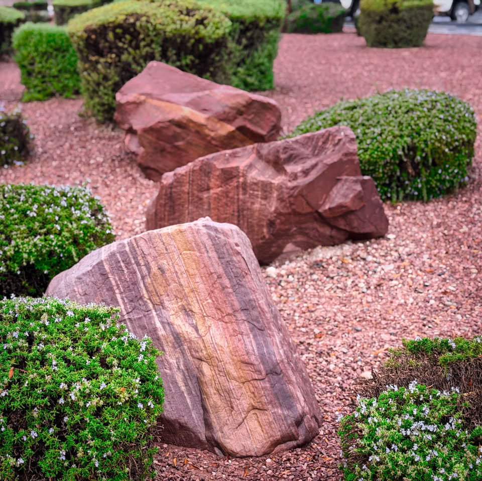 A landscaped garden with large red rocks and neatly trimmed green bushes on a bed of red gravel.