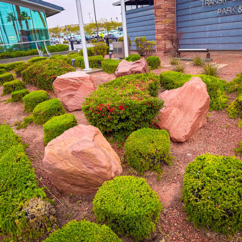 Landscaped garden with large red rocks and neatly trimmed green shrubs, adjacent to a building with glass windows and a brick wall.