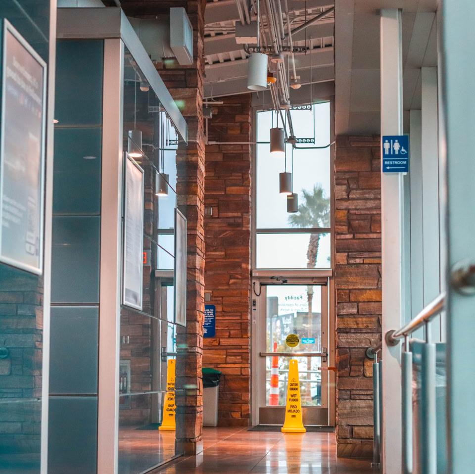 A modern hallway with glass walls, a polished floor, and a brick wall at the end, featuring large windows and overhead lights.