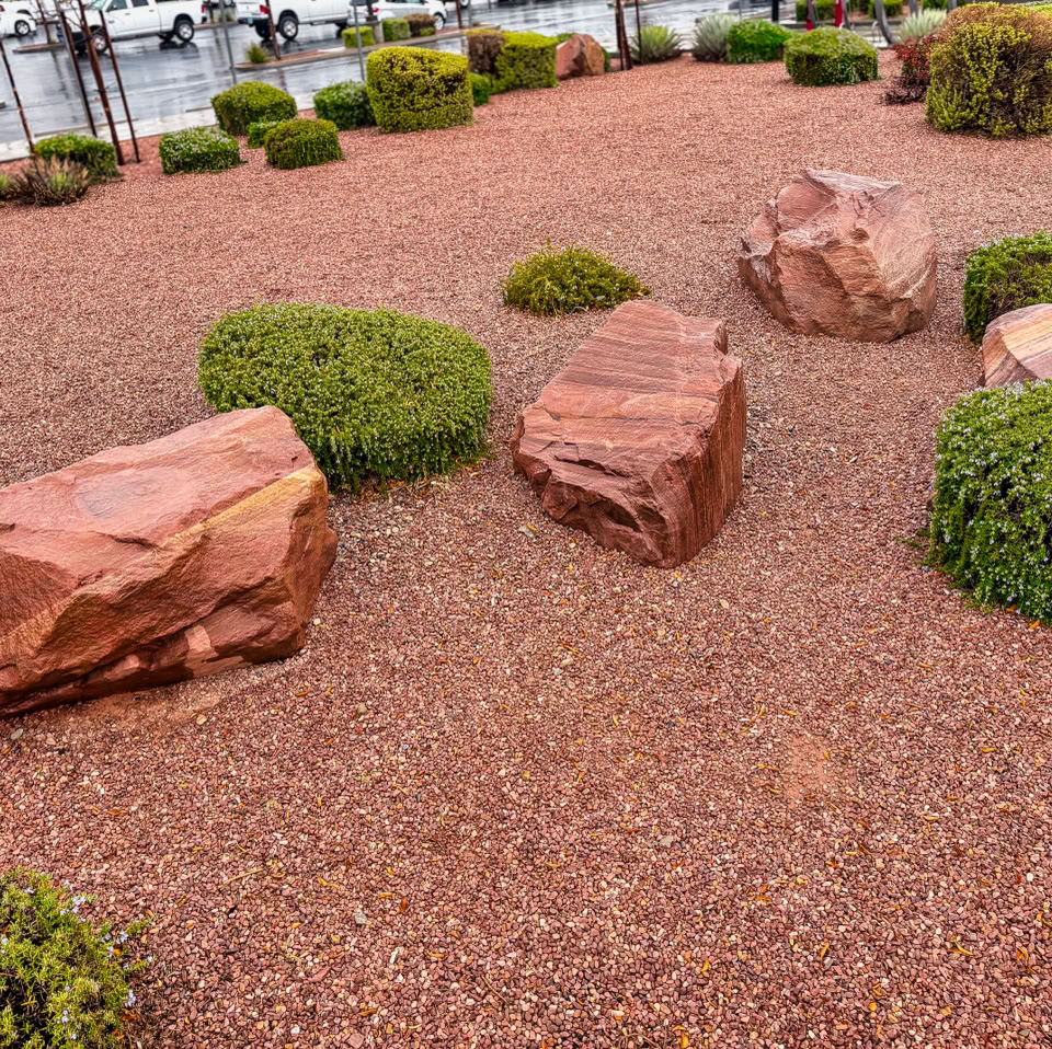 A landscaped area with red rocks and green shrubs on a gravel surface.