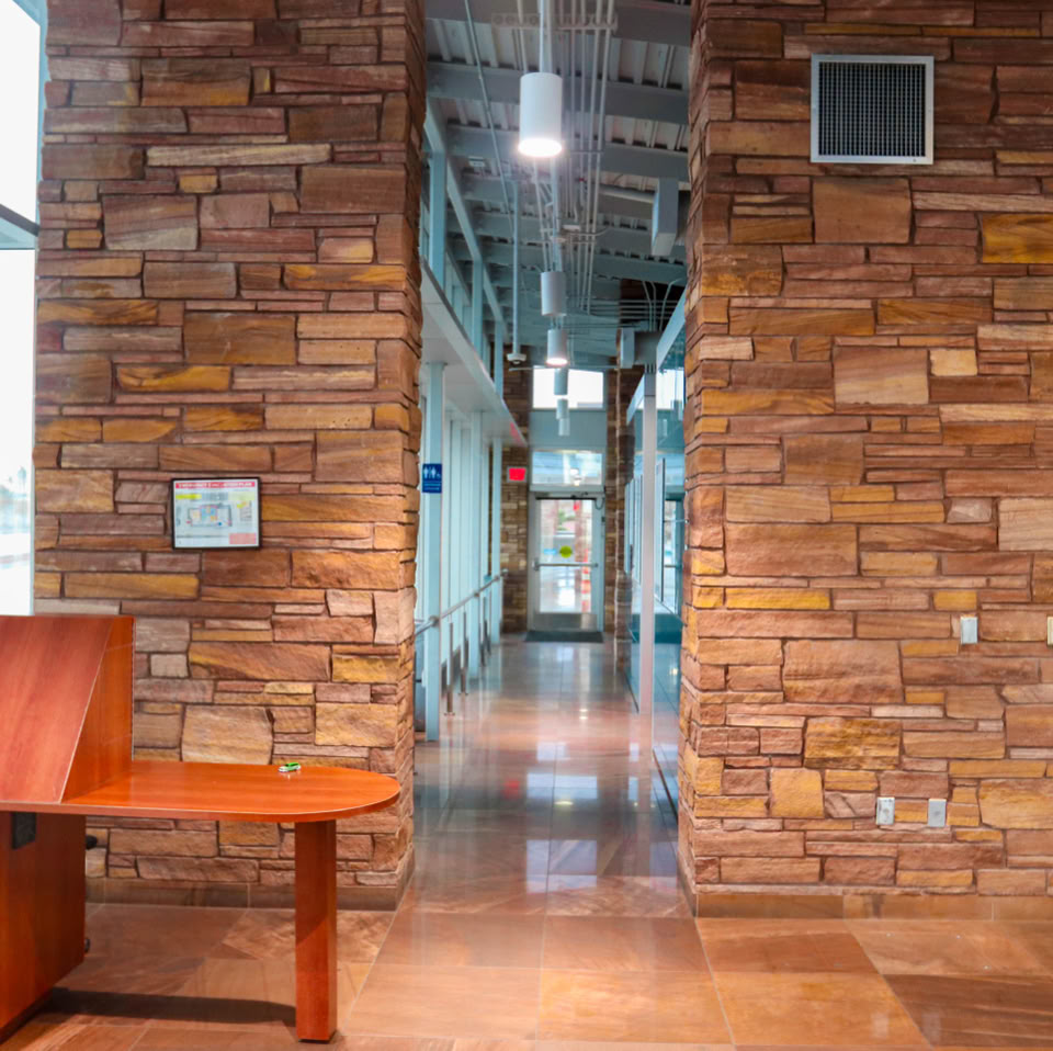 A hallway with stone walls, a wooden desk on the left, and large windows on the left side.