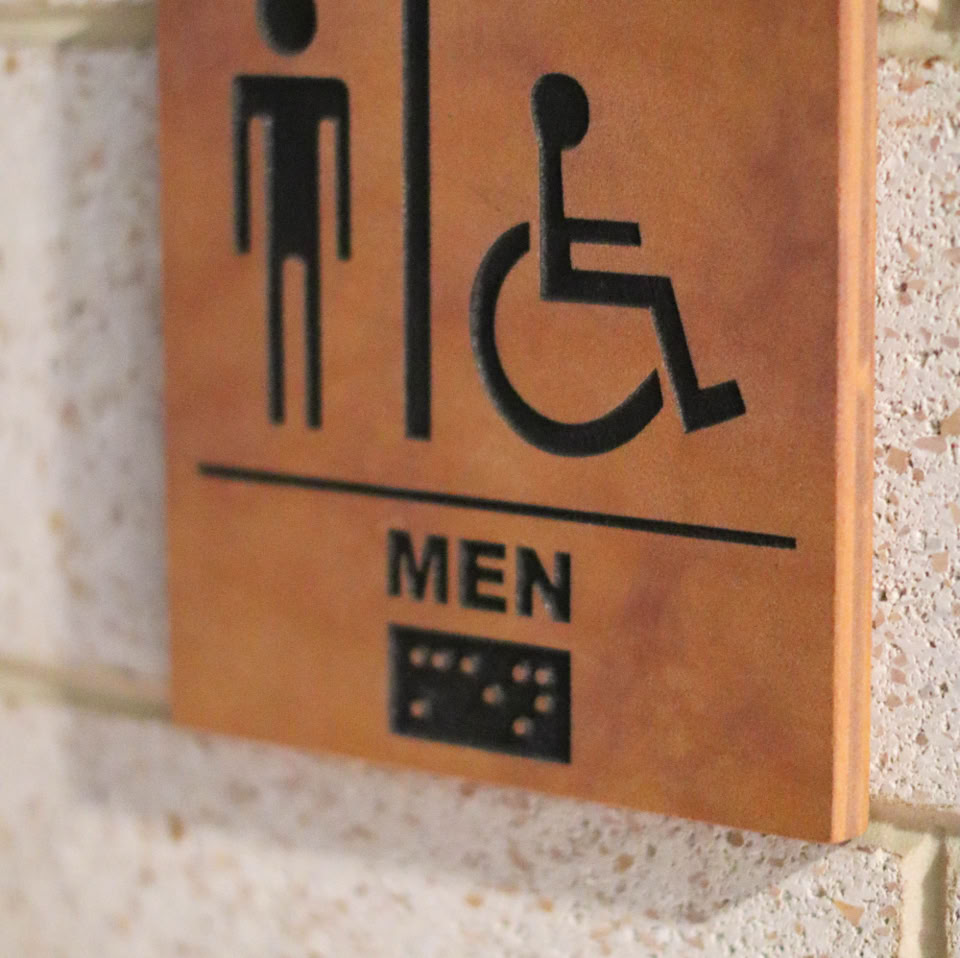 A wall-mounted sign indicating a men's restroom with wheelchair accessibility, featuring male and wheelchair symbols, the word "MEN," and Braille text.