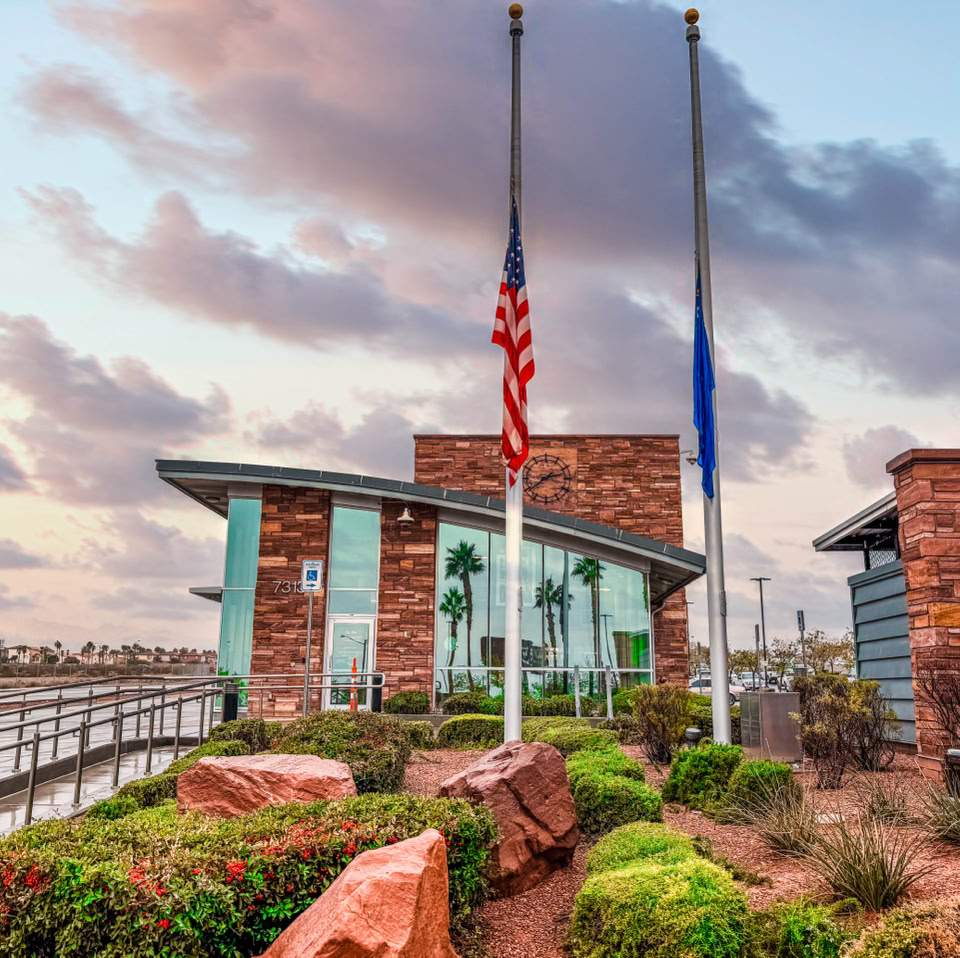 Modern building with large glass windows, two flagpoles with flags at half-mast, surrounded by landscaped greenery and red rocks, under a cloudy sky.
