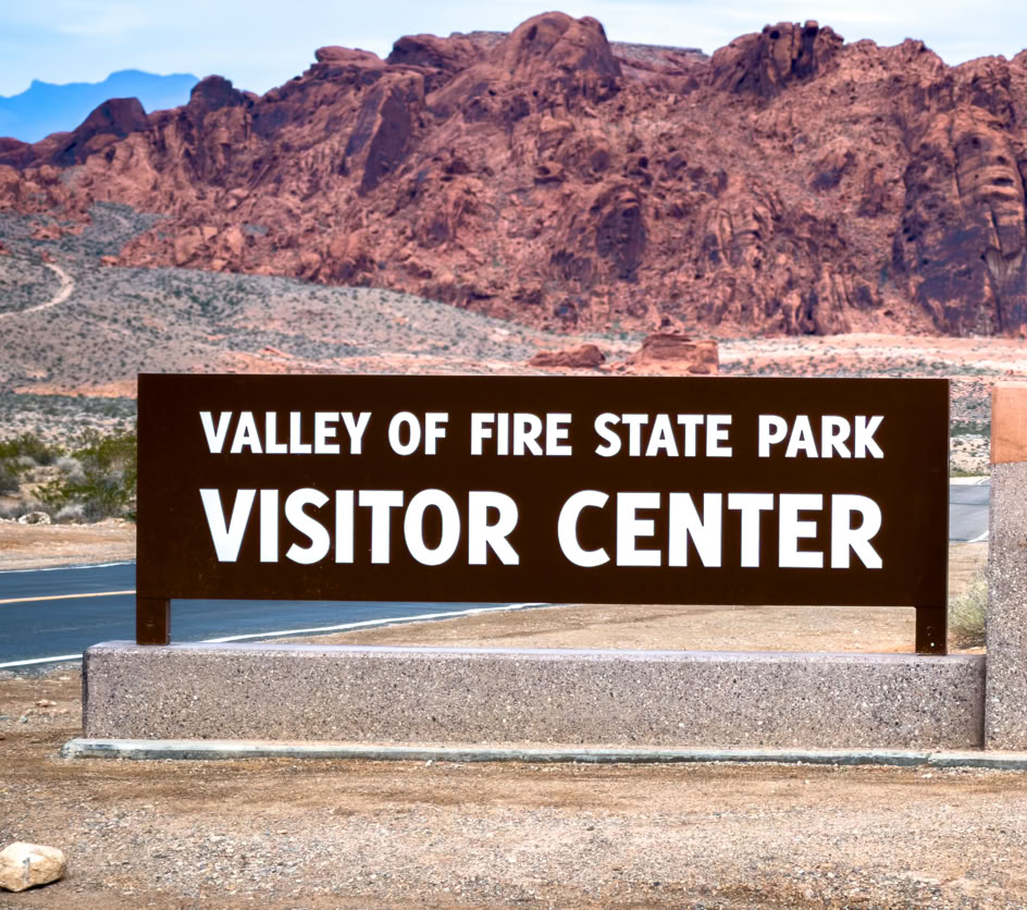 Sign reading "Valley of Fire State Park Visitor Center" with red rock formations in the background.