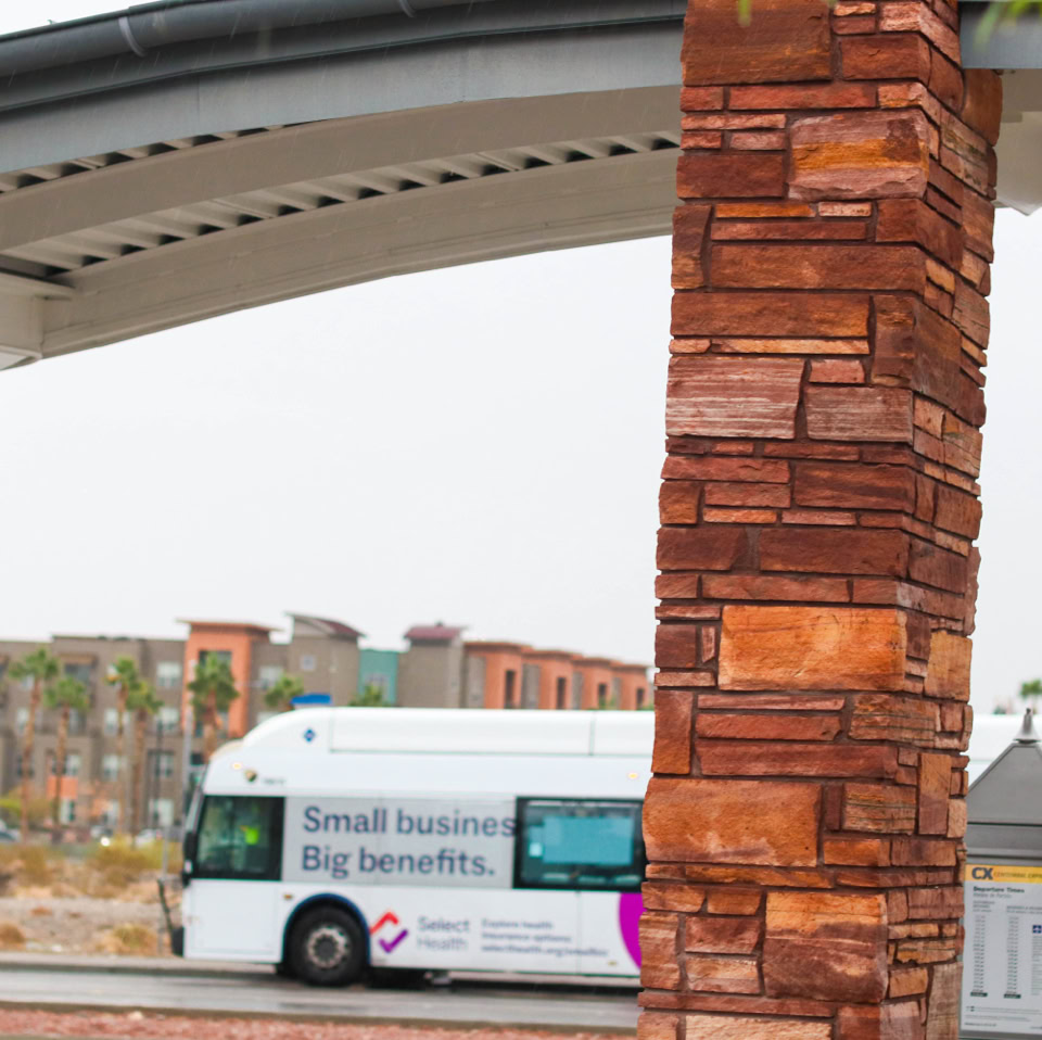 A white bus with text on its side is parked near a brick pillar under a curved structure. In the background, there are buildings and palm trees.