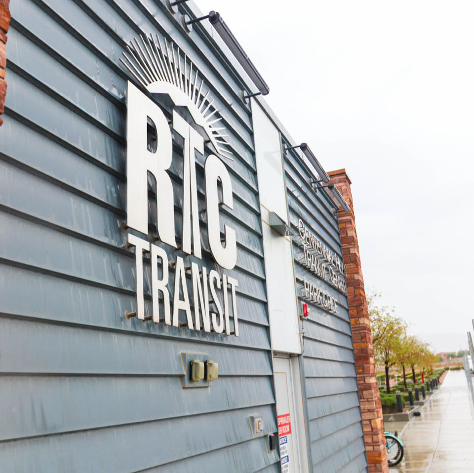 Exterior of a building with a sign reading "RTC Transit" on a wall with horizontal panels, adjacent to a brick section, with a parking lot and trees in the background.