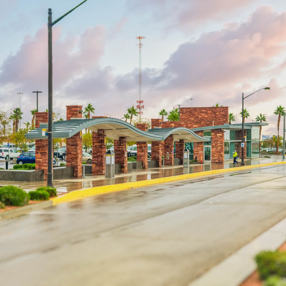 A street scene with a covered bus stop featuring brick columns and a wavy roof, surrounded by greenery, with a cloudy sky in the background.