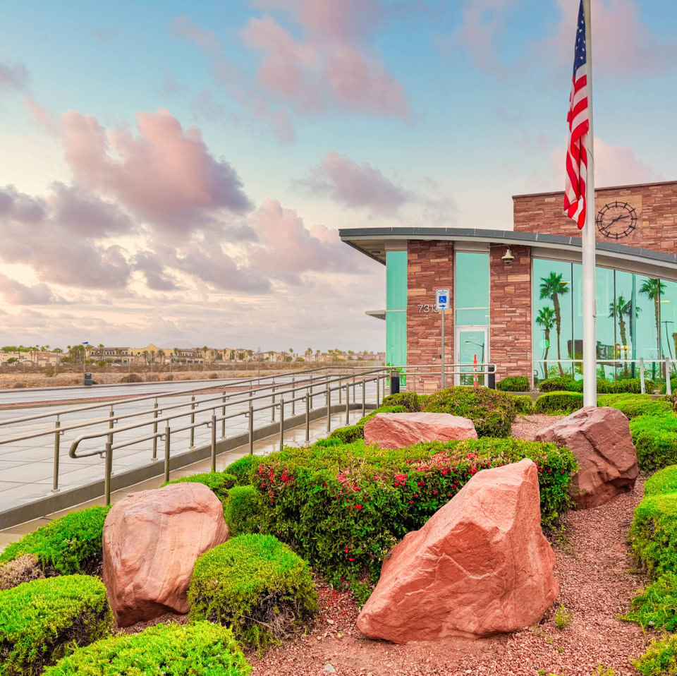 A modern building with large glass windows and a brick facade, surrounded by landscaped greenery and rocks. Two flagpoles with flags are in front, and a cloudy sky is in the background.