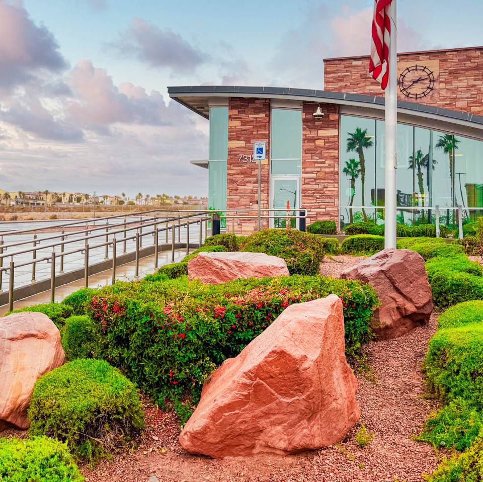 Modern building with large glass windows and stone facade, surrounded by landscaped greenery and red rocks, with flagpoles in the foreground.