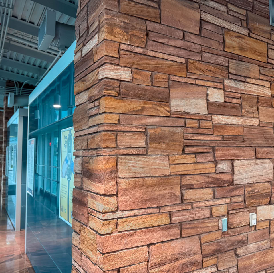 Interior view of a modern building with a stone wall on the right and glass windows on the left, featuring exposed ceiling pipes and polished floors.
