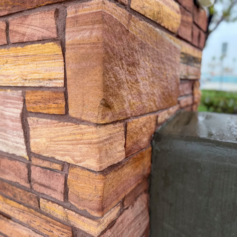 Close-up of a corner of a brick wall with a textured, reddish-brown pattern, adjacent to a smooth, gray surface. Trees and sky are blurred in the background.