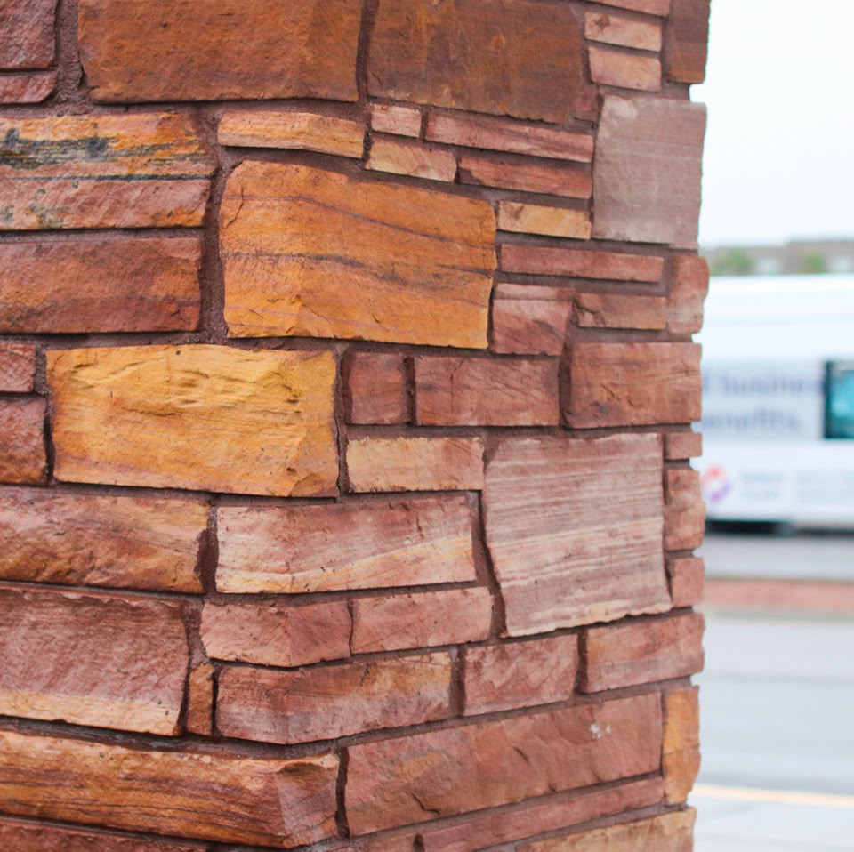 A close-up of a stone pillar with rectangular and irregularly shaped stones in various shades of brown and red. In the background, a blurred view of a street and a vehicle with a purple and blue design.