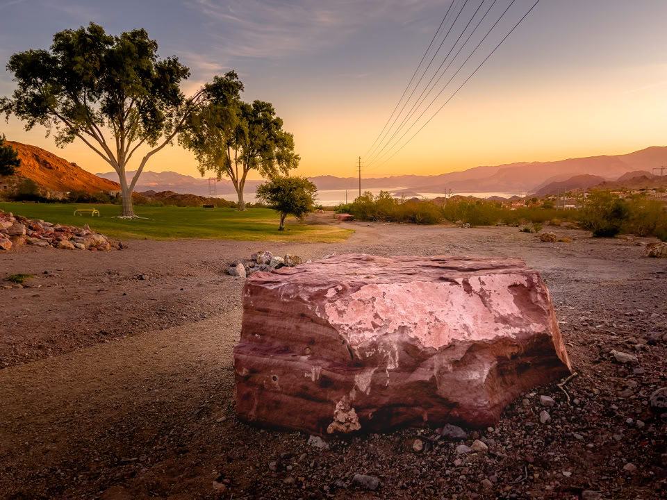 A large pink rock on a dirt path with trees, power lines, and a sunset over distant hills and water in the background.