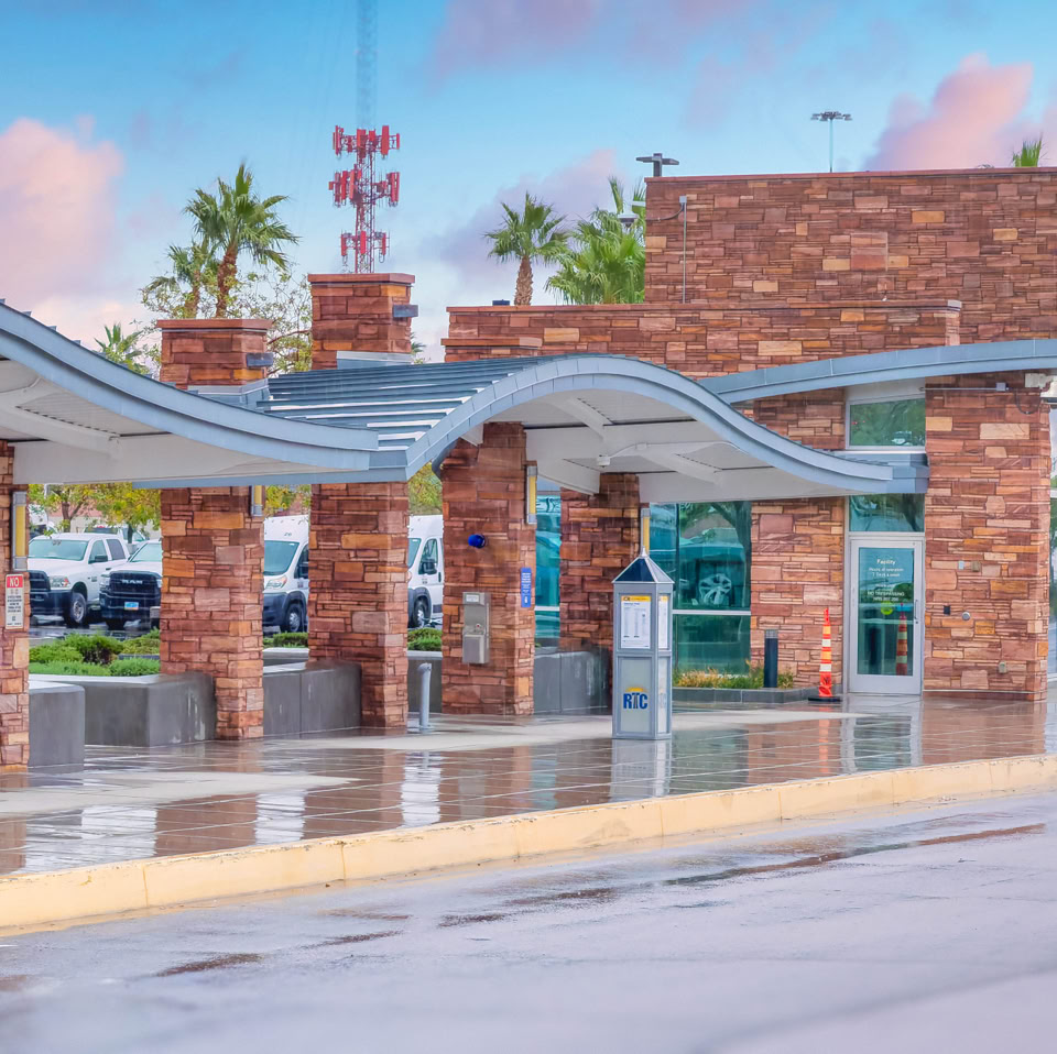 A modern outdoor structure with wavy metal roofing supported by brick columns, set against a backdrop of parked cars and palm trees under a blue sky with clouds.