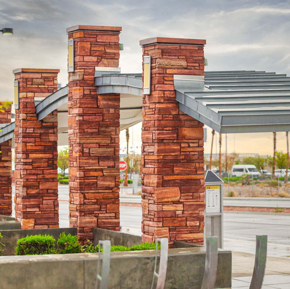 Brick columns supporting a metal canopy structure near a road, with greenery and a cloudy sky in the background.