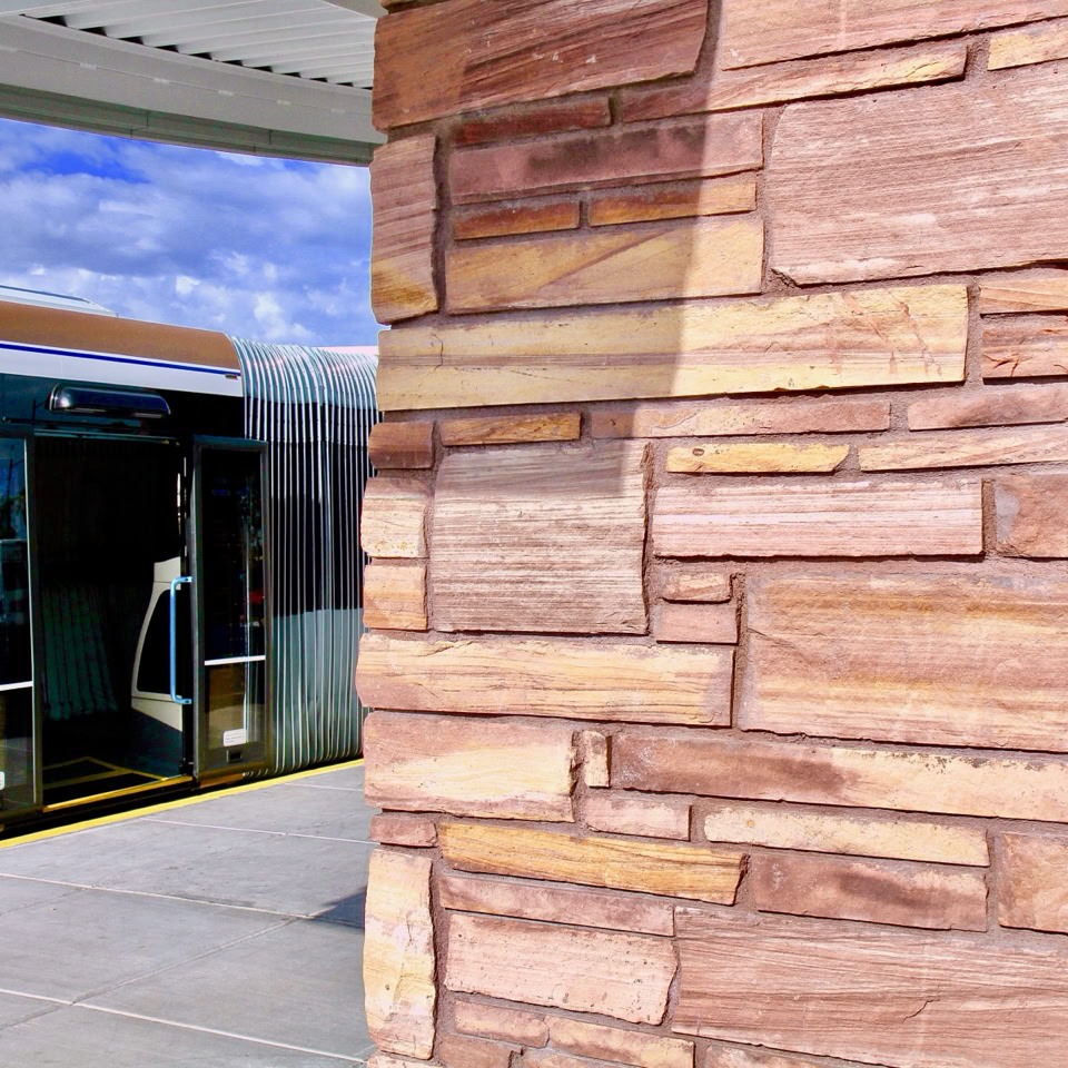 A stone pillar in the foreground with a modern train station and train in the background under a blue sky.