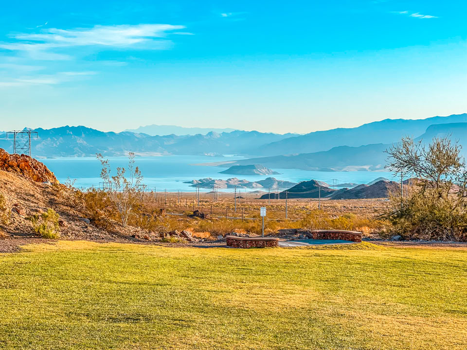 A scenic landscape with a grassy foreground, sparse shrubs, and a view of a lake surrounded by hills under a clear blue sky HEMENWAY PARK.