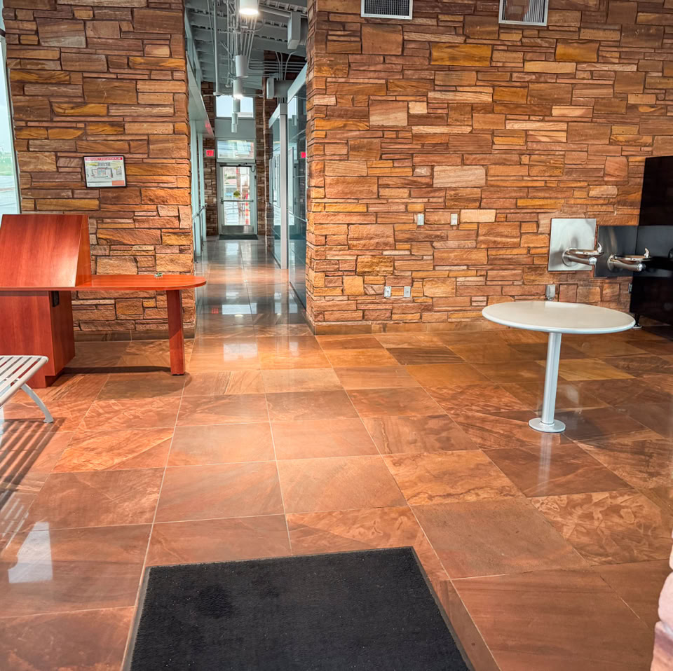 Interior view of a room with stone walls and a shiny tiled floor. There is a wooden bench on the left, a round table in the center, and a desk with a computer on the right.