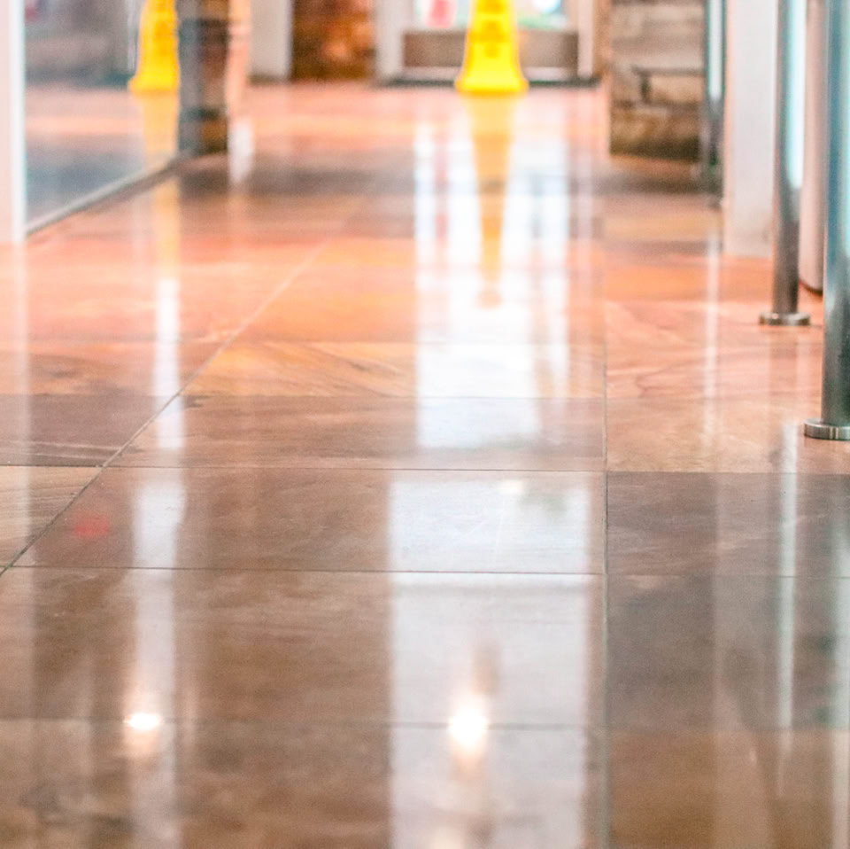 A shiny, reflective hallway floor with metal railings on the right and blurred yellow caution signs in the background.