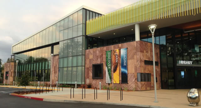 A modern building with large glass windows and a Stone facade, featuring banners and a sign that reads "Library" near the entrance. at Mitchell Park Library